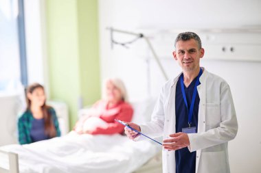 İyi aydınlatılmış bir hastane odasında bir kadın ve ailesiyle konuşurken bir doktor elinde bir pano ile ayakta duruyor. Bu sahne hastaya destek ve bakım sağlar..