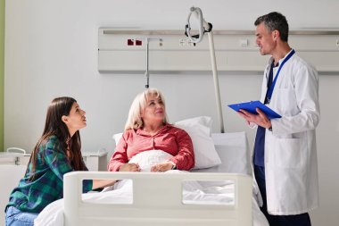 Aile üyeleri dikkatle dinlerken bir doktor, hasta yatağında yatarken bakım planlarını tartışır. Ayar, destekleyici bir atmosfer yaratan, parlak bir hastane odası..