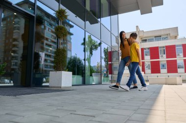 Bir kadın ve genç bir çocuk şehir merkezinde geziniyor. Modern binalara yakınlar, güneşli bir günün tadını çıkarıyorlar. Ağaçlar ve ekiciler sahneye doğanın bir dokunuşunu ekler..