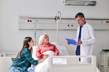 Bir doktor hastane odasında bir hastayla sağlık güncellemelerini tartışırken, bir ziyaretçi destek önerir. Olay yeri tıbbi bir ortamda bakım ve endişeyi gösteriyor..