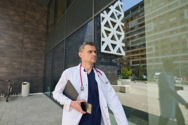 Steteskoplu bir sağlık uzmanı modern bir tıbbi tesisten ayrılıyor. Dizüstü bilgisayar taşıyor ve odaklanmış görünüyor. Şehir batımında güneş parlıyor..