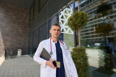 Çağdaş bir tıp binasının önünde elinde telefon ve not panosu olan bir sağlık uzmanı duruyor. Arkasında modern mimari ile odaklanmış ve meşgul görünüyor..