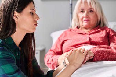 Genç bir bakıcı, bir hastane yatağında yaşlı bir kadının elini nazikçe tutarken, parlak ve temiz bir sağlık hizmetine yapılan samimi bir ziyaret sırasında sıcaklığı ve şefkati iletirken görülüyor..