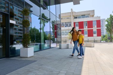 Bir anne ve oğlu çağdaş bir binanın önünde kaldırımda yürüyorlar. Birlikte güneşli bir günün tadını çıkarıyorlar. Bölgeyi keşfederken bir ışık anı paylaşıyorlar..