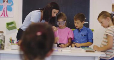 İlkokul öğrencilerine fen sınıfında deney tüpleri ve sıvılarla kimya deneyi yoluyla güvenlik gözlüğü takan bir öğrenciye rehberlik eden kadın öğretmen. Sinema yavaşlığı