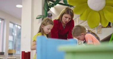 Gülümseyen bayan öğretmen iki mutlu ilkokul kızına çizimleri ve boyalarıyla yardımcı olmak için bir sınıfta yaratıcı sanat dersi sırasında masaya eğiliyor..