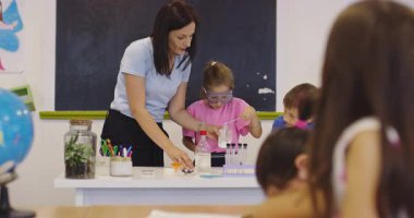 İlkokul öğrencilerine fen sınıfında deney tüpleri ve sıvılarla kimya deneyi yoluyla güvenlik gözlüğü takan bir öğrenciye rehberlik eden kadın öğretmen. Sinema yavaşlığı