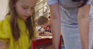 Parlak bir anaokulunda resim dersi sırasında renkli kalemlerle resim çizen yaratıcı küçük kızlara yardım eden bir bayan öğretmen..