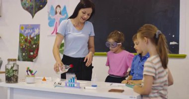 İlkokul öğrencilerine fen sınıfında deney tüpleri ve sıvılarla kimya deneyi yoluyla güvenlik gözlüğü takan bir öğrenciye rehberlik eden kadın öğretmen. Sinema yavaşlığı