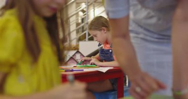 Parlak bir anaokulunda resim dersi sırasında renkli kalemlerle resim çizen yaratıcı küçük kızlara yardım eden bir bayan öğretmen..