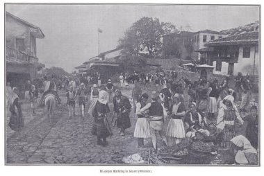 Geleneksel elbiseli insanların, meyve ve mal satan satıcıların ve tarihi bir Arnavut kasabasında hareketli bir kalabalığın bulunduğu kaldırımlı bir sokakta hareketli bir pazar sahnesi..