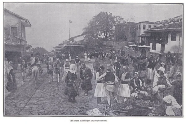Geleneksel elbiseli insanların, meyve ve mal satan satıcıların ve tarihi bir Arnavut kasabasında hareketli bir kalabalığın bulunduğu kaldırımlı bir sokakta hareketli bir pazar sahnesi..