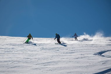 Üç kayakçı güneşli, toz kaplı bir dağ yamacını oyup açık mavi gökyüzünün altında taze kar çıkararak dinamik bir kış sporu sahnesi yaratıyor..