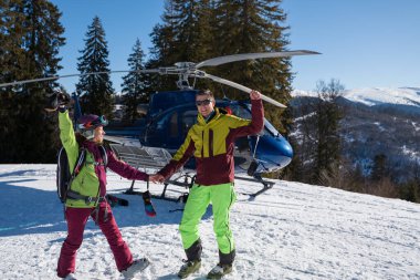 Parlak kayak kıyafetleri içinde gülümseyen bir adam ve kadın, güneşli dağlarda helikopter yolculuğunu kutlarken mavi bir helikopterin yanında karlı bir arazide duruyorlar..