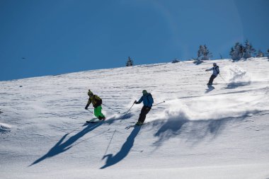 Üç kayakçı güneşli, toz kaplı bir dağ yamacını oyup açık mavi gökyüzünün altında taze kar çıkararak dinamik bir kış sporu sahnesi yaratıyor..