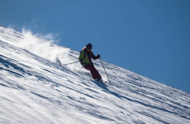 Dişi bir kayakçı güneşli bir dağ yamacında taze tozu oyar, inerken karı tekmeler, açık mavi gökyüzü altında dinamik ve enerjik bir kış sporu sahnesi yaratır..