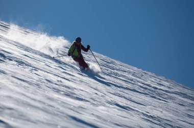 Dişi bir kayakçı güneşli bir dağ yamacında taze tozu oyar, inerken karı tekmeler, açık mavi gökyüzü altında dinamik ve enerjik bir kış sporu sahnesi yaratır..