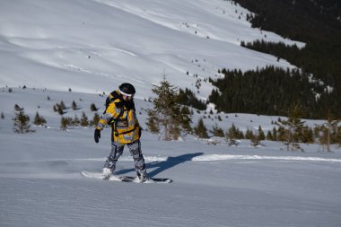 Sarı ve gri kamuflaj kıyafetli bir snowboardcu güneşli bir kış gününde el değmemiş bir kar yamacından manzaralı bir dağ ormanı geçmişiyle iniyor..