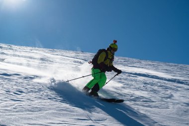 Güneşli bir dağ yamacında kayak yapan kayakçı, o inerken kar tekmeliyor, açık mavi gökyüzü altında dinamik ve enerjik bir kış sporu sahnesi yaratıyor..