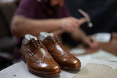Cilalanmış kahverengi deri ayakkabılar çalışma masasında dinlenirken, el ve aletler arka planda bulanıklaşıyor, bir zanaatkârın dükkan sahnesini yakalıyor ve detaylara dikkat çekiyor..