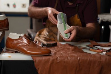 Önlüklü bir zanaatkar, işlek bir atölyede kahverengi deri parçaları ve ayakkabı yapma aletleriyle çevrili bir mezura ile deri taban ölçüsü alır..
