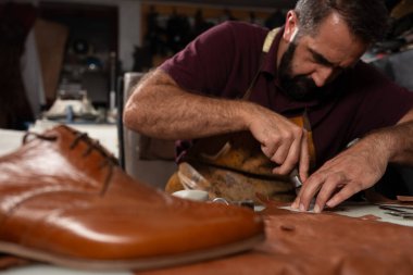 Odaklanmış bir deri işçisi dağınık bir atölyede kahverengi deri parçalarını buduyor. Ön planda, el işçiliğini ve titiz deri işçiliğini ön plana çıkaran cilalı bir ayakkabı var..