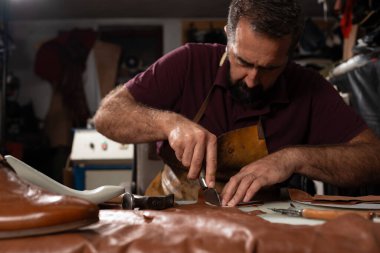 Yetenekli bir deri ustası, loş bir atölyede, deri keserek ve şekillendirerek dikkatle çalışır. Deri bir önlük giyiyor ve kemer, çanta ve diğer parçaları yapmak için bıçak ve aletler kullanıyor..