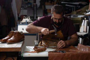 Yetenekli bir zanaatkar kahverengi deriyi şekillendirmeye ve dikmeye yoğunlaşır, dağınık bir atölyede çekle ve ağızla. Bitmiş ayakkabılar, aletler ve deri kırıntıları masayı doldururken yakınlarda oturuyor..