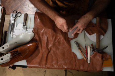 Deri bir zanaatkar, ayakkabı yapma araçları, desenler ve derilerle çevrili yoğun bir çalışma masasında deri bir parça dikiyor. Sahne, geleneksel zanaat, hassasiyet ve el işçiliğini yakalıyor..
