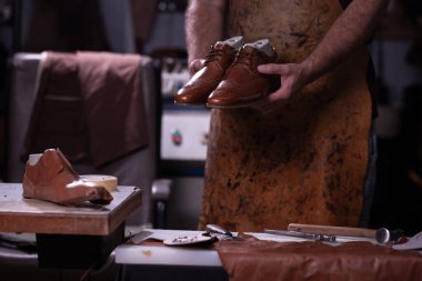 Lekeli önlüklü bir zanaatkar meşgul bir ayakkabı yapma atölyesinde bir çift kahverengi deri ayakkabı sunar. Aletler ve tamamlanmamış deri parçalar masanın üzerinde, zanaatkarlığı yakalıyor..