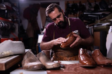 Sakallı yetenekli bir ayakkabıcı yoğun bir atölyede kahverengi deri bir ayakkabıyı inceler. Birkaç bitmiş ve tamamlanmamış çift, geleneksel ayakkabı imalatı ve dikkatli el işçiliğinin altını çizerek onu çevreledi..