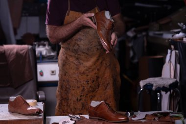 Lekeli önlük giyen bir zanaatkar, dağınık bir atölyede aletler ve diğer ayakkabılarla çevrili kahverengi bir deri ayakkabıyla çalışır. El işçiliğini ve detaylara dikkat çekmeyi vurgular..