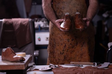 Lekeli önlüklü yetenekli bir zanaatkar, dağınık bir ayakkabı yapma tezgahı, bant ve deri parçaları, zanaatkar zanaat ve onarım ve onarım arasında bir çift cilalı kahverengi deri ayakkabı sunar..