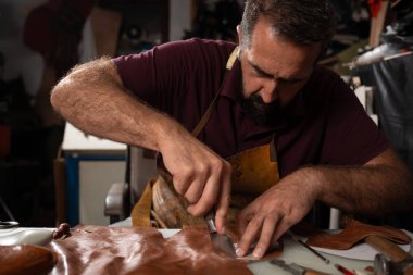 Deri önlük giyen sakallı bir zanaatkar, loş bir atölyede, kahverengi deriyi bir aletle kesiyor, kemer üzerinde çalışıyor. İşçiliğe odaklanırken etrafını aletler ve kırıntılar sarmış..