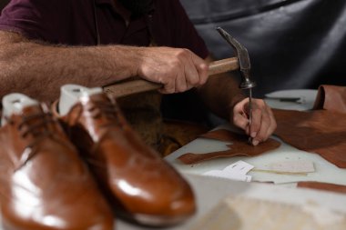 Yetenekli bir deri ustası ayakkabı için deri parçaları şekillendirmek için çekiç kullanır. Kalıp kalıntıları ve araçlar çalışma tablosunun üzerinde yatarken cilalanmış kahverengi ayakkabılar yakınlarda dinleniyor.