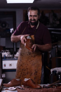 Yetenekli bir ayakkabıcı dağınık bir atölyede duruyor, elinde parlak kahverengi bir ayakkabı var. Lekeli deri bir önlük giyiyor ve gülümsüyor. Etrafı aletlerle ve bitmiş ayakkabılarla çevrili. Müşteriler için hazır..