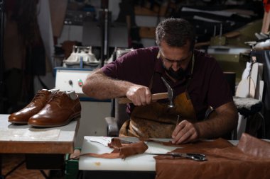 Bir zanaatkar tezgahta oturur ve kahverengi ayakkabıları birleştirmek için deri parçaları çekiçler. Deriler ve aletlerle çevrelenmiş, odaklanmış yapıcı, yetenek, sabır ve adanmışlık gösteriyor