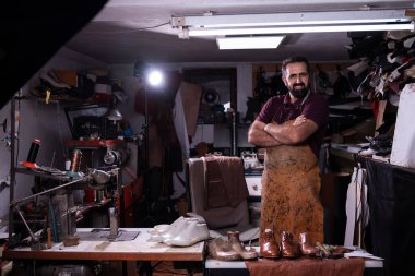 Lekeli önlüklü bir zanaatkar, etrafı aletlerle, dikiş makinesiyle çevrili, dağınık bir deri atölyesinde kollarını kavuşturup el işçiliğiyle gurur duyan, bitirilmiş ayakkabılar giyer..