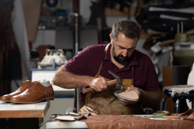 Yetenekli bir deri işçisi dağınık bir stüdyoda ayakkabı şekillendirmeye konsantre olur. Yoğun atölye araçlar, deri kırıntıları ve bitmiş bir çift, işçilik ve dikkat ile dolu