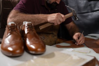 Yetenekli bir ayakkabıcı, bir çekiç ve keski kullanarak bir tezgah üzerinde deri şekillendirirken, bir çift cilalı kahverengi ayakkabı ön planda duruyor. El işi, işçilik ve deri işleri tam ekrana sunuldu..