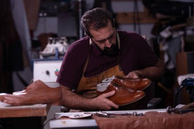 Önlük giymiş yetenekli bir zanaatkar, malzeme ve aletlerle dolu dağınık bir atölyenin içinde ayrıntıları inceleyen parlak kahverengi deri ayakkabılar taşır. Kaliteli, odaklı ve el yapımı