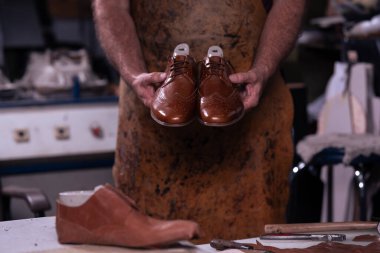 Lekeli önlüklü yetenekli bir zanaatkar, işlek bir atölyede parlak kahverengi deri ayakkabılar, daha fazla ayakkabı ve araç gereç, nakliye aracı, onarım ve zanaat kalitesinde sunar..