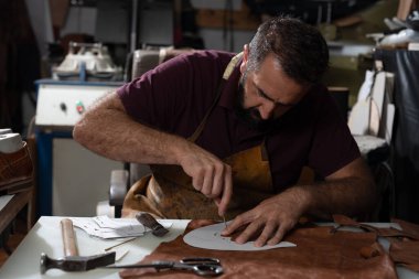 Odaklanmış bir deri işçisi, önlük giyerken keskin bir aletle kahverengi deriyi şekillendirir. Çeşitli aletler ve kırıntılar bankı dolduruyor, geleneksel el sanatlarını yakalıyor ve işlek bir deri stüdyosunun havasını yakalıyor..