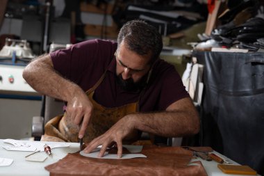 Yetenekli bir deri işçisi dağınık bir masada oturur ve dikkatlice deri bir parçayı desen boyunca keser. Aletler, kırıntılar ve yıpranmış bir önlük, yoğun bir atölyede geleneksel, pratik işçiliği ön plana çıkarıyor..