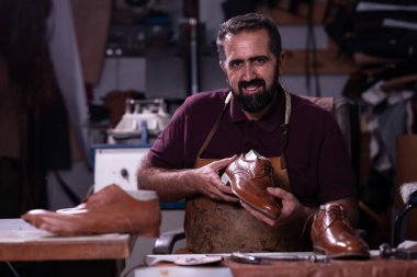 Sakallı ve önlüklü yetenekli bir zanaatkar hareketli bir atölyede kahverengi deri bir ayakkabı sunar. Aletler ve deri parçalarla çevrili halde, el yapımı ayakkabı işçiliği sergiliyor..