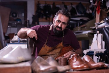Önlük giymiş yetenekli bir sakallı zanaatkar dağınık bir atölyede kahverengi deri ayakkabılar üzerinde çalışıyor, el yapımı deri kaplama ve geleneksel ayakkabı yapımı..