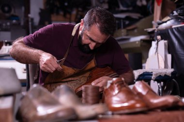 Sakallı bir zanaatkar cilalı deri bir ayakkabı üzerinde çalışır, iyi aletler ve deri bir önlük kullanır. Ayakkabılar ve malzemeler onu yoğun bir atölyede çevrelemiş, el işçiliği ve dikkat çekiyor