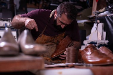 Deri önlük giyen bir zanaatkar, diğer ayakkabılar ve atölyede iş aletleriyle çevrili kahverengi deri bir ayakkabıyı teftiş eder. Sahnede hassasiyet, ustalık ve geleneksel sanatlar vurgulanıyor.