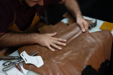 Bir zanaatkar, etrafını kesme aletleri ve dikiş kalıplarıyla çevrili büyük bir kahverengi deri parçasını pürüzsüz hale getirir. Sahne, işçilik, odaklanma ve el yapımı deri ürünler sanatını yansıtıyor..