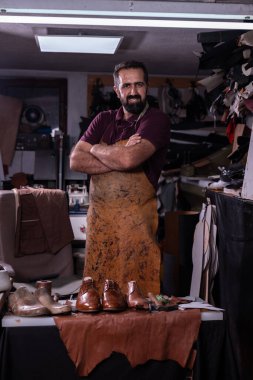 Becerikli bir deri ustası dağınık atölyesinde poz veriyor, üzerinde lekeli bir önlük var ve beş cilalı ayakkabı masanın üzerinde duruyor. Etrafını saran aletler, kırıntılar ve deri derileri işçiliğin altını çiziyor.